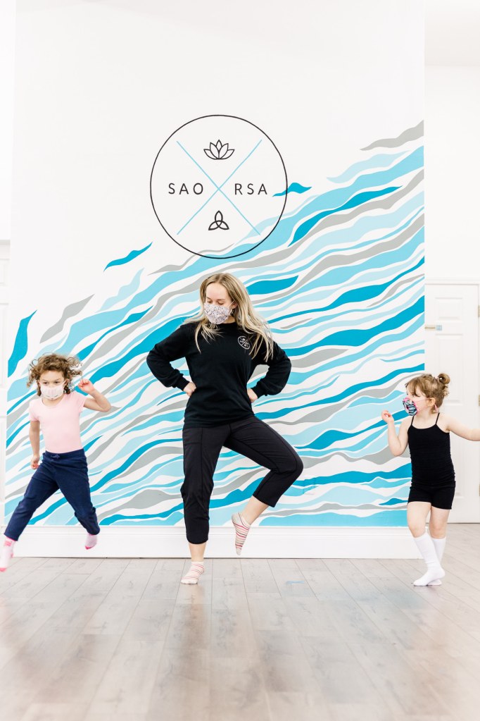 A woman and two children jump in front of a blue wall that reads "SAORSA"