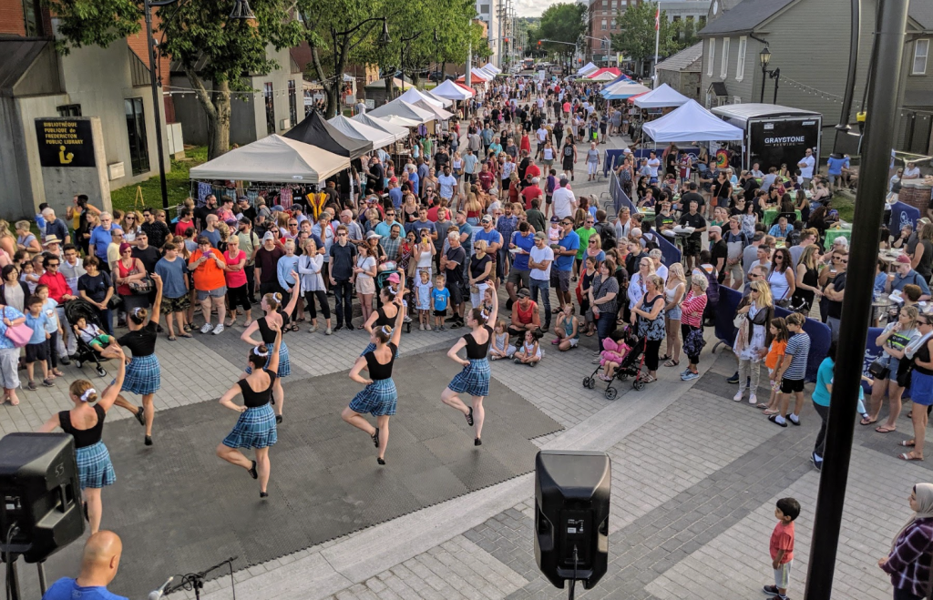 A city street is filled with a crowd watching a group of 7 dancers in blue kilts perform on a black stage