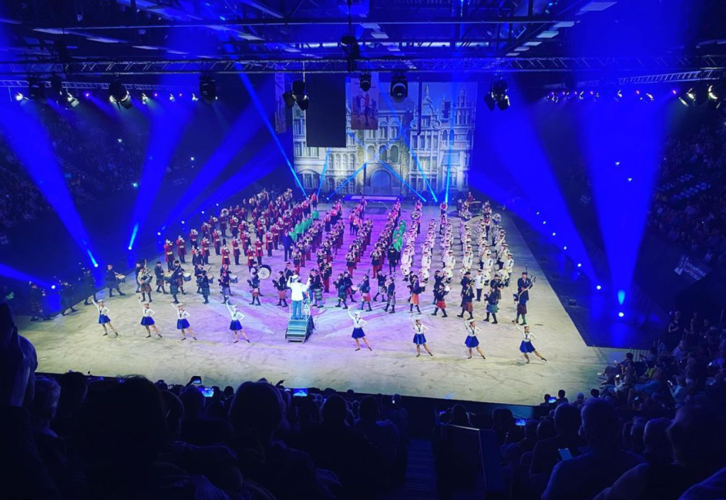 A large cast of performers in lines in an arena with crowds on either sides and purple lighting