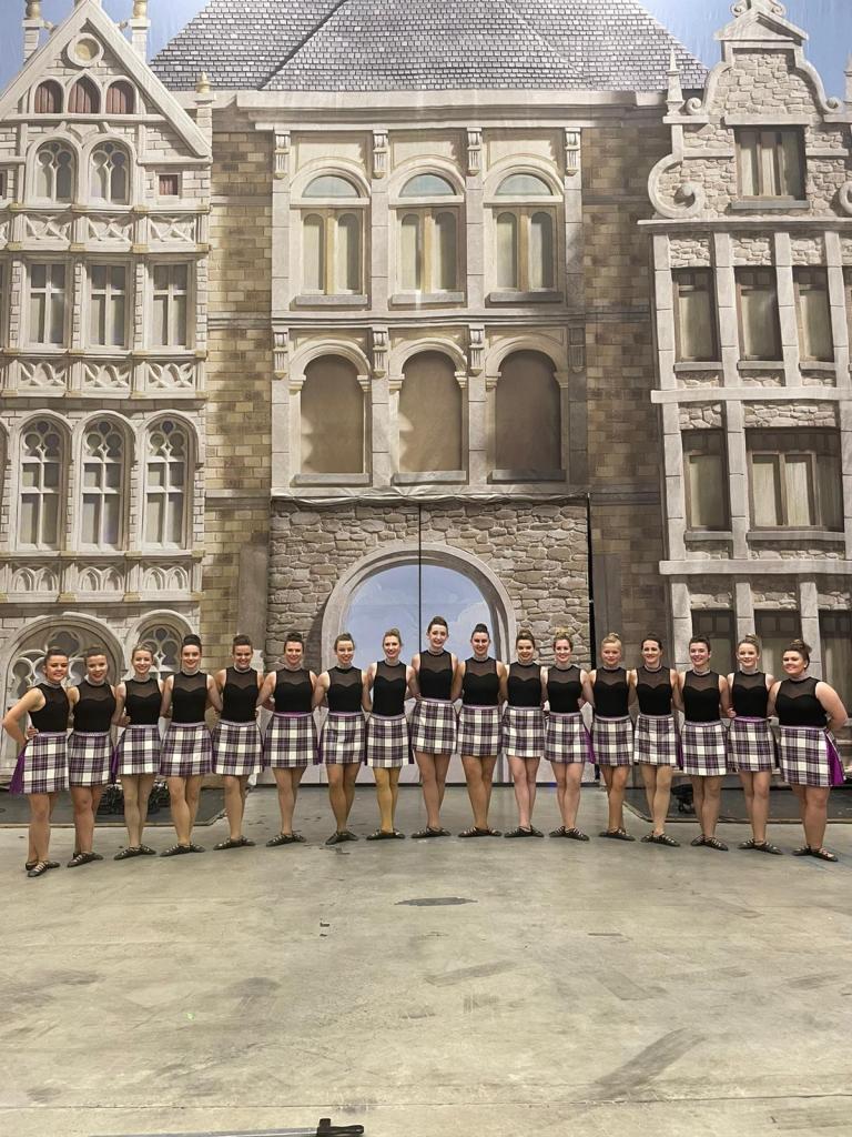 A link of dancers in black tops and purple kilts in front of a castle