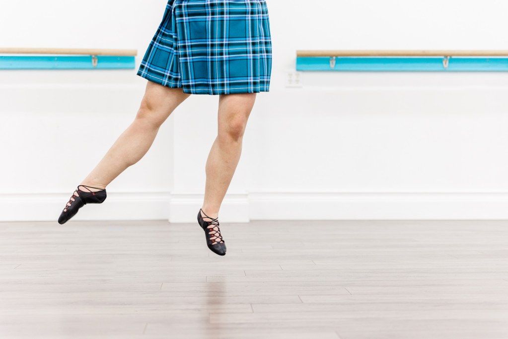 A dancer's legs as they jump in a white room wearing black dance shoes and a blue kilt