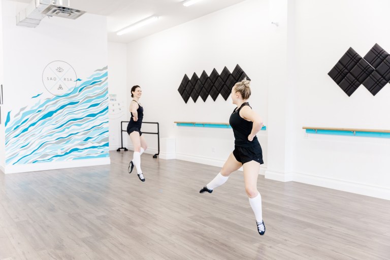 Two dancers jumping in a white room with a blue wall that reads "SAORSA"