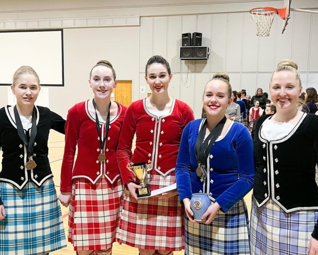 5 dancers in multi colour kilts smile with medals and trophies