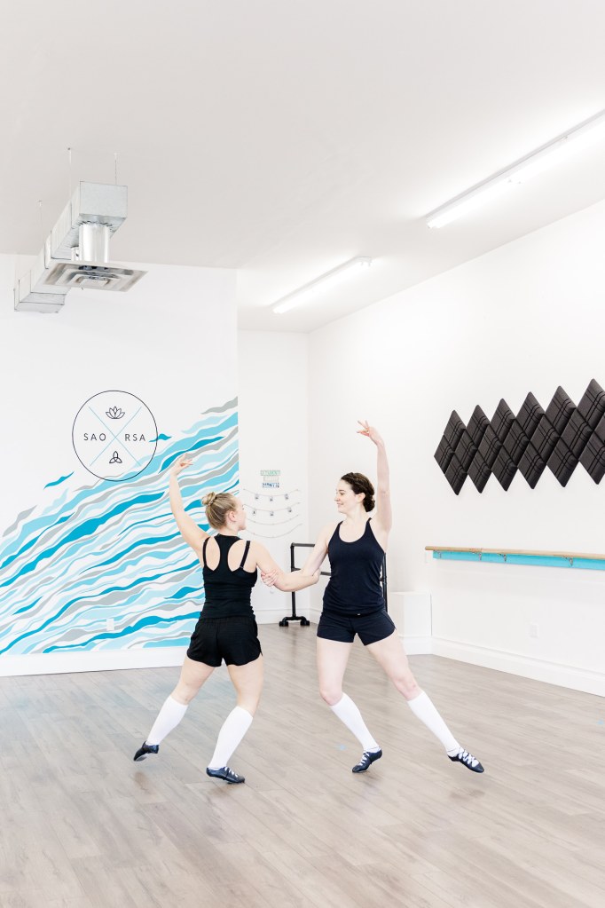 Two dancers together in a white room with a blue wall that reads "SAORSA"