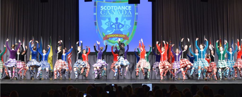 Many dancers in multi colour kilts dance on a black stage