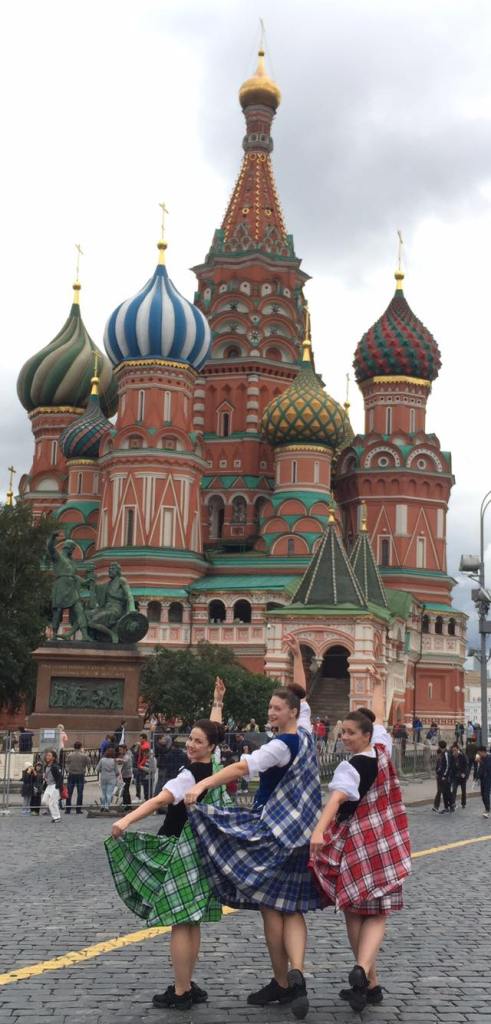 Three dancers in plaid outfits pose in front of a castle