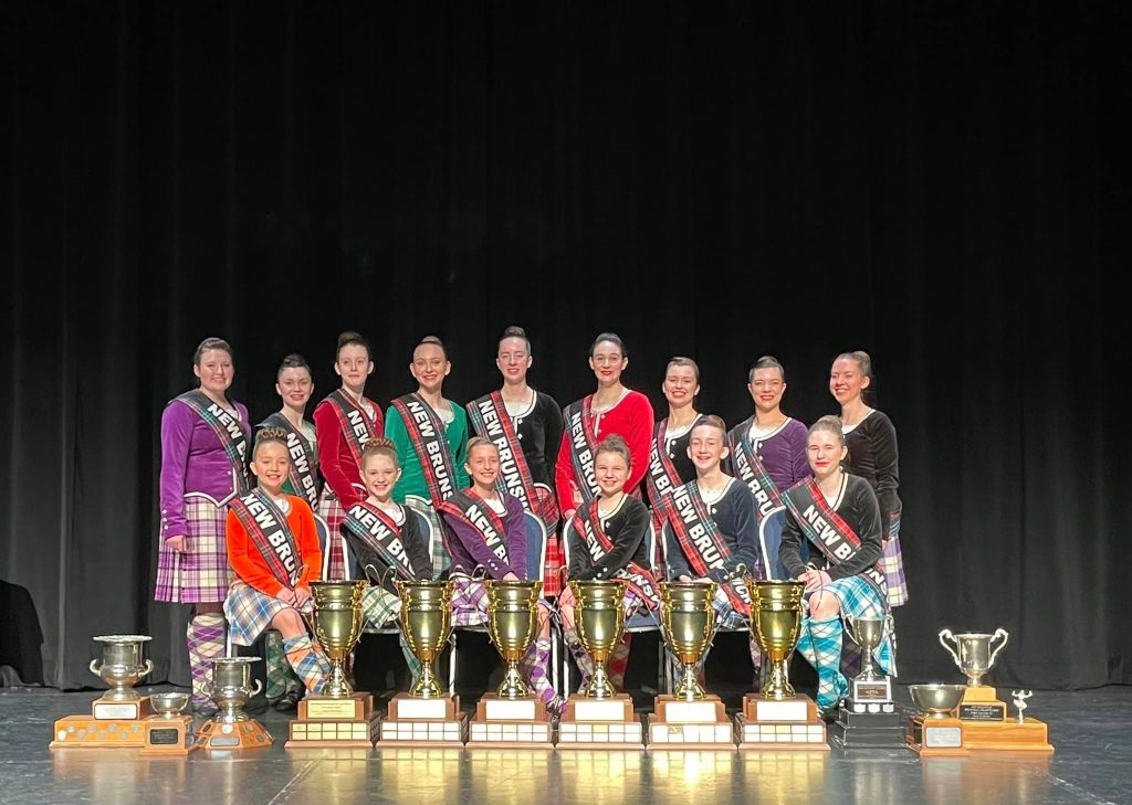 A group of dancers in multi colour kilts smile with trophies