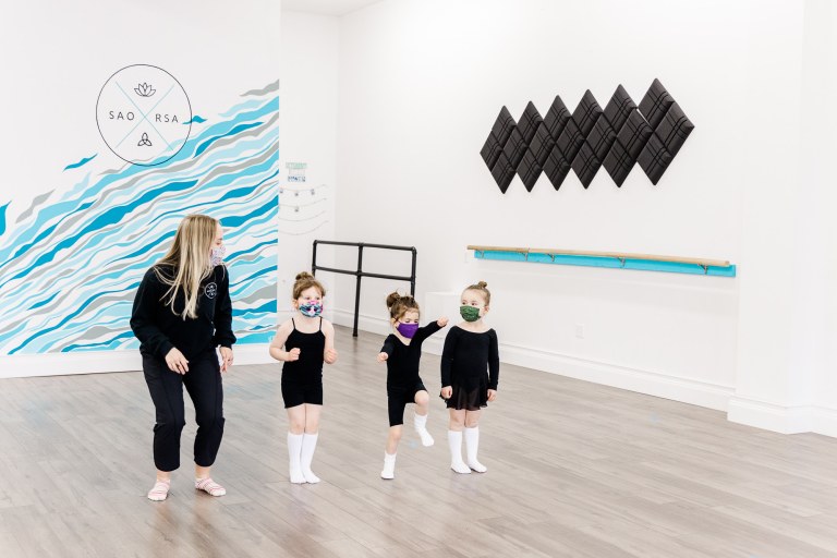 Three dancers and an instructor wearing black in a white room with a blue wall that reads "SAORSA"