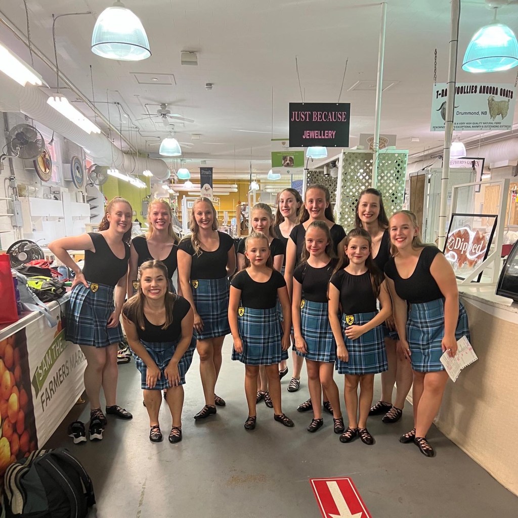 A large group of people wearing black tops and blue kilts smile