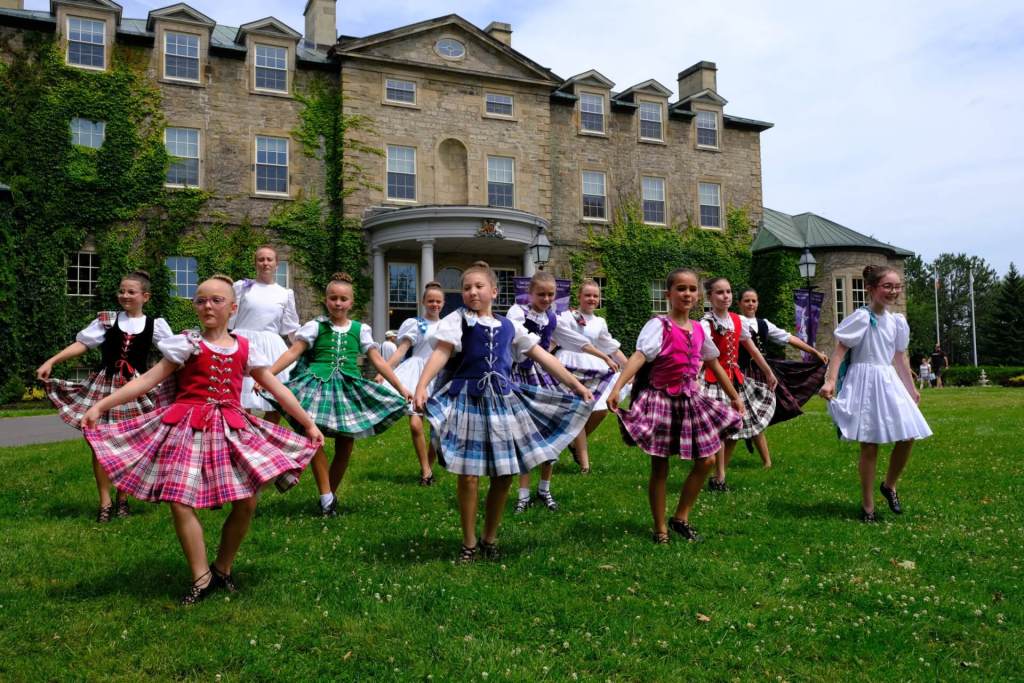 A group of many young girls dancing in plaid outfits in front of a stone mansion