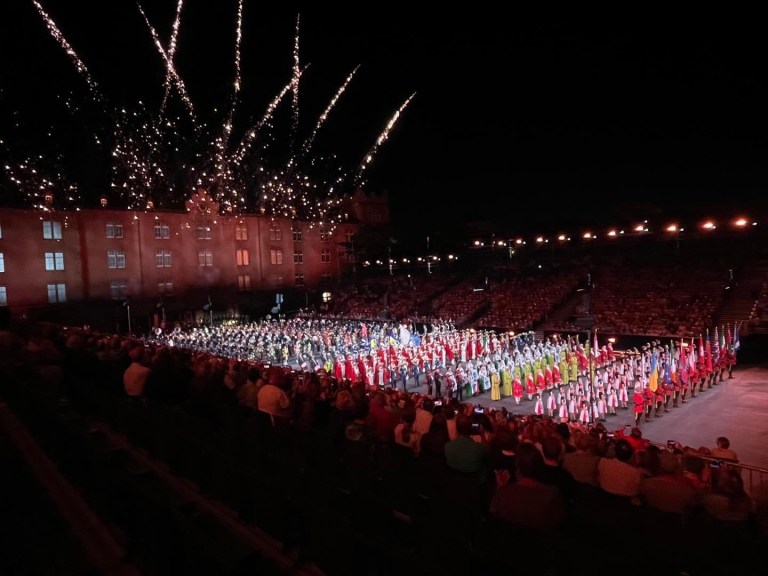 A large arena with a crowd, many performers on a large stage and fireworks in the background