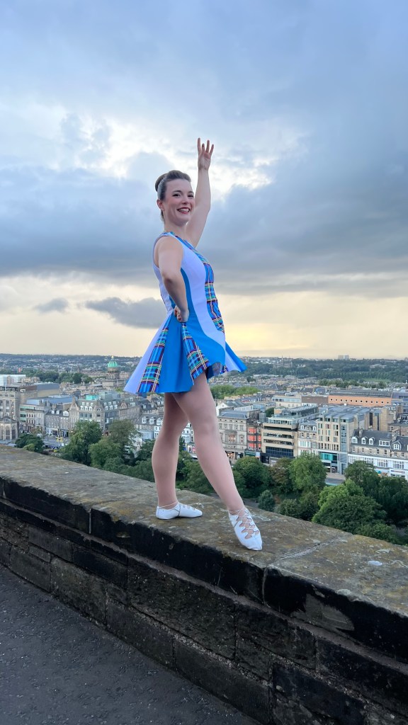 A woman in a blue dress stands posed on a balcony overlooking Edinburgh
