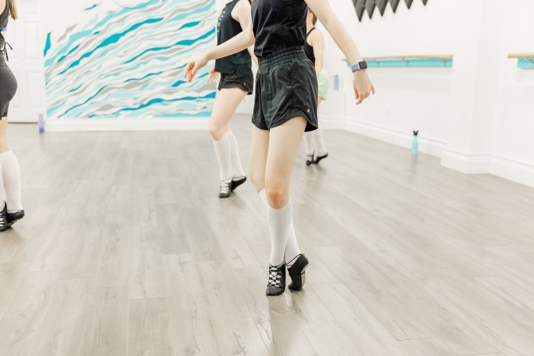 4 Dancers posed on their toes wearing white knee socks and black dance shoes in a white room