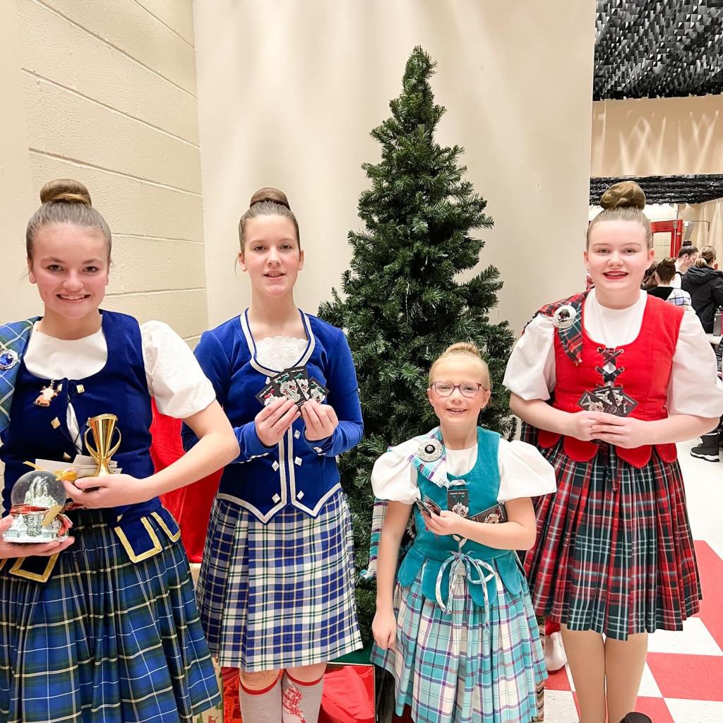 4 dancers in kilts smile in front of a christmas tree