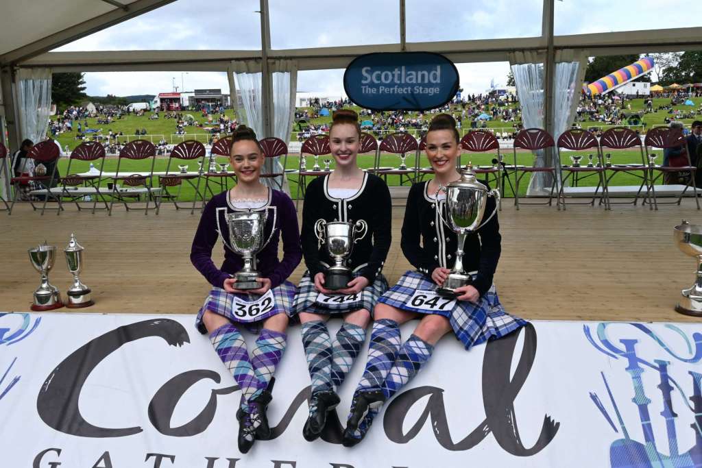 Three dancers smile holding trophies sitting on a stage that reads "Cowal"