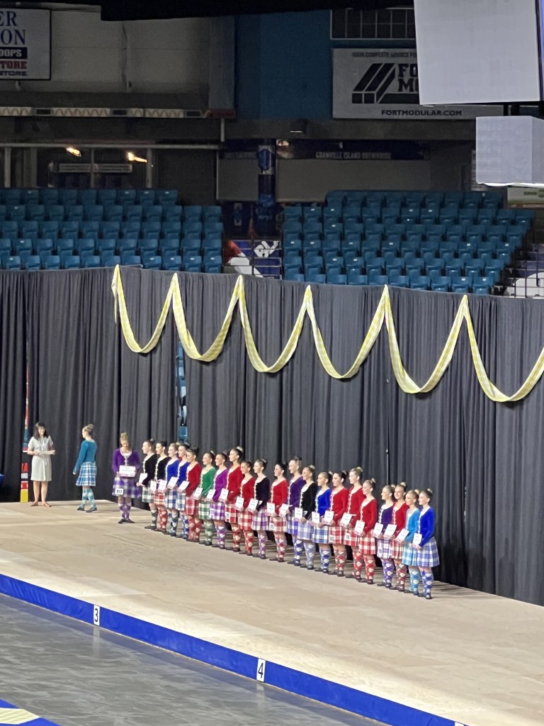 A line of dancers in a kilt stand holding certificates on a stage