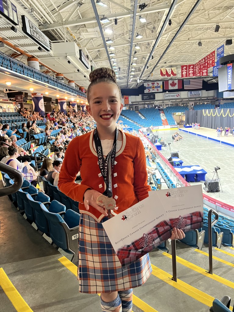 A dancer in an orange kilt smiles holding a certificate