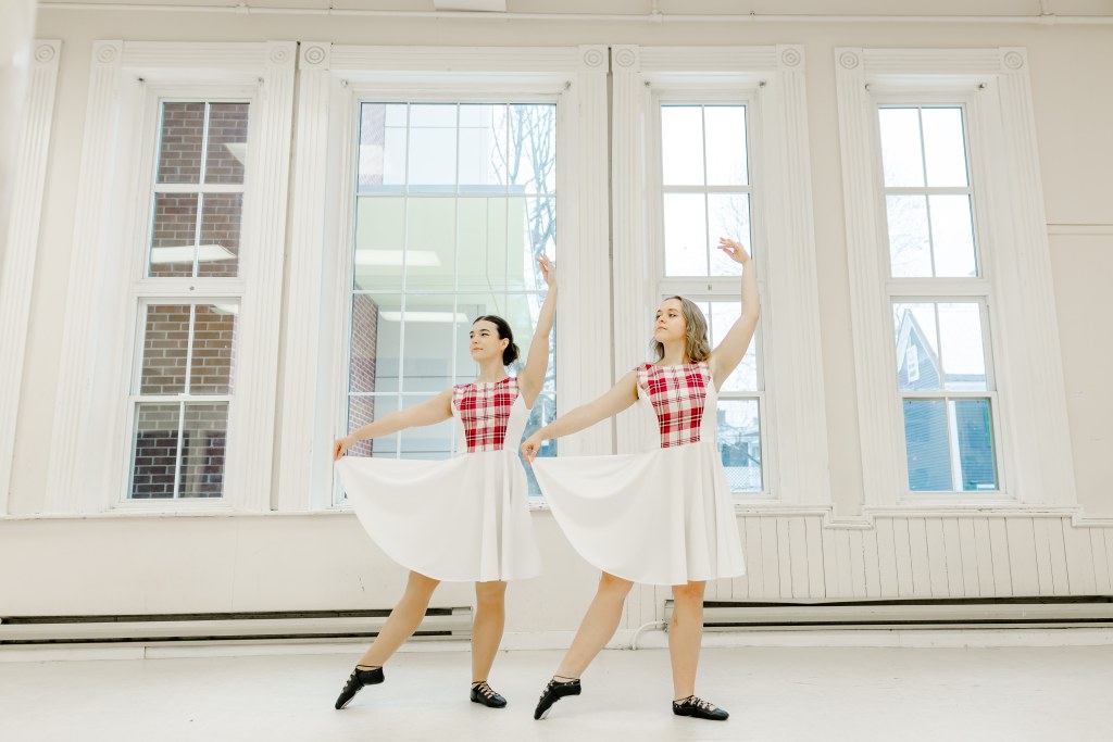 Two dancers in white and pink dresses pose in front of large windows