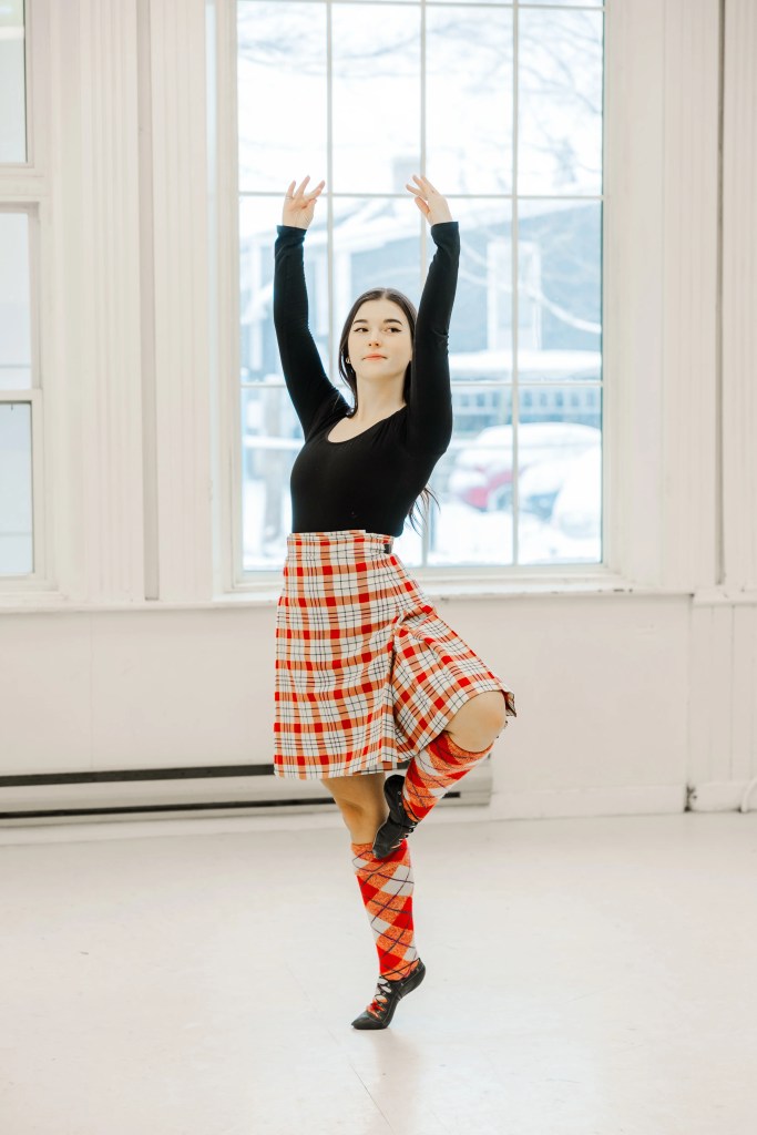 A highland dancer wearing a red kilt outfit demonstrates a movement in a white studio. Saorsa Studio offers Highland Dance classes for ages 1 to adult in Fredericton, New Brunswick.