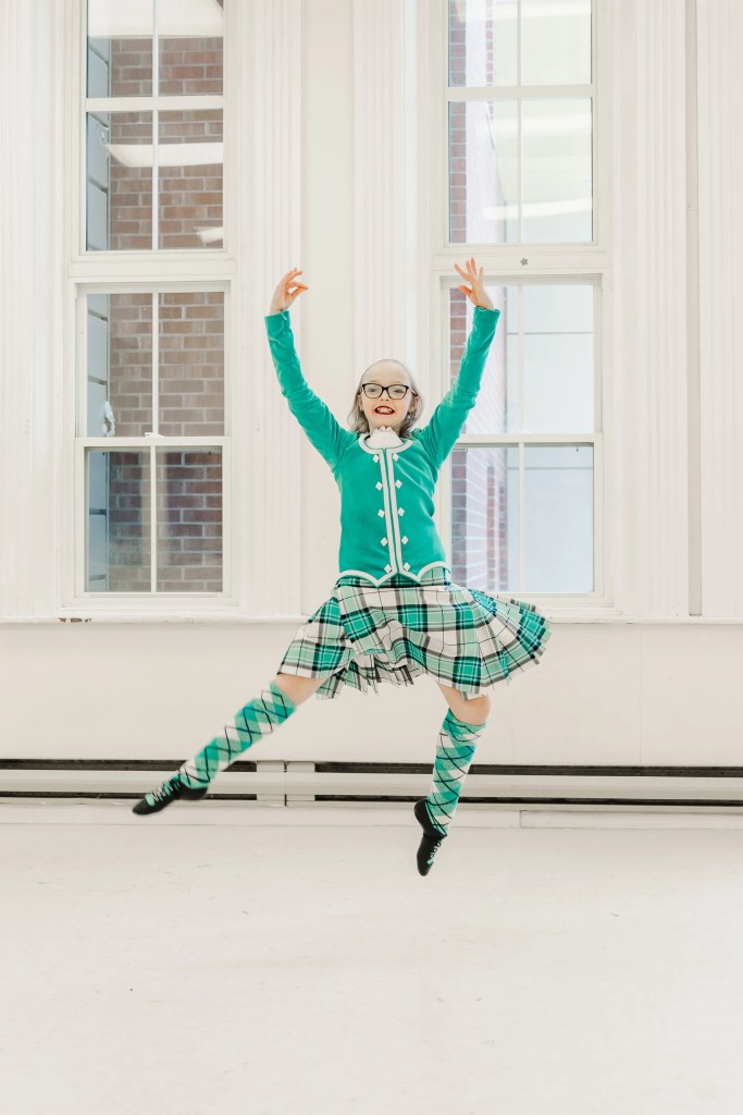A highland dancer wearing a teal kilt outfit demonstrates a movement in a white studio. Saorsa Studio offers Highland Dance classes for ages 1 to adult in Fredericton, New Brunswick.