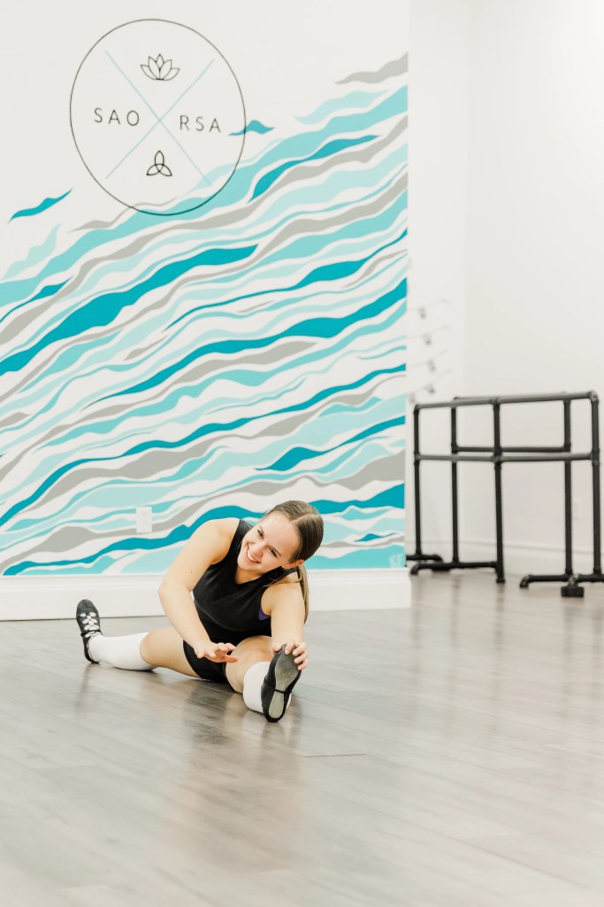 A highland dancer, Kate Francoeur, sits on the ground stretching. The wall behind her is white and blue and reads "Saorsa". Saorsa Studio offers Highland Dance classes for ages 1 to adult in Fredericton, New Brunswick.