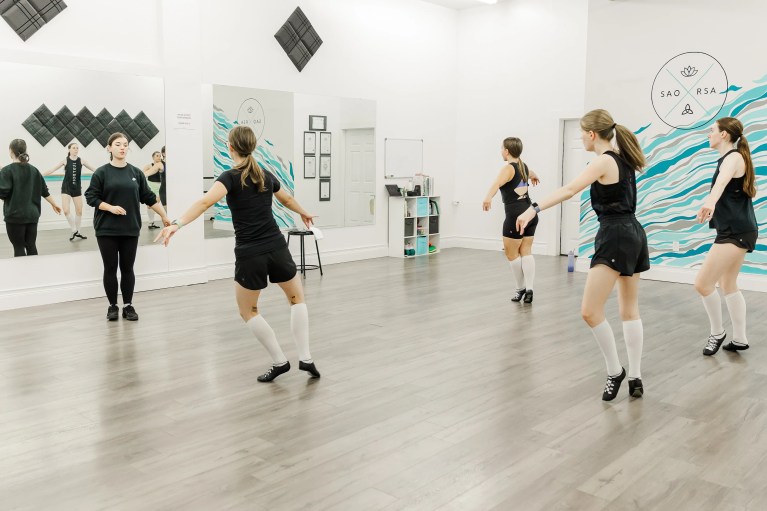 Highland dancers wearing black, with white socks and black leather dance ghillies shoes. They stand poised on the balls of their feet. There is an instructor in all black at the front of the room. The studio space is white and blue. Saorsa Studio offers Highland Dance classes for ages 1 to adult in Fredericton, New Brunswick.