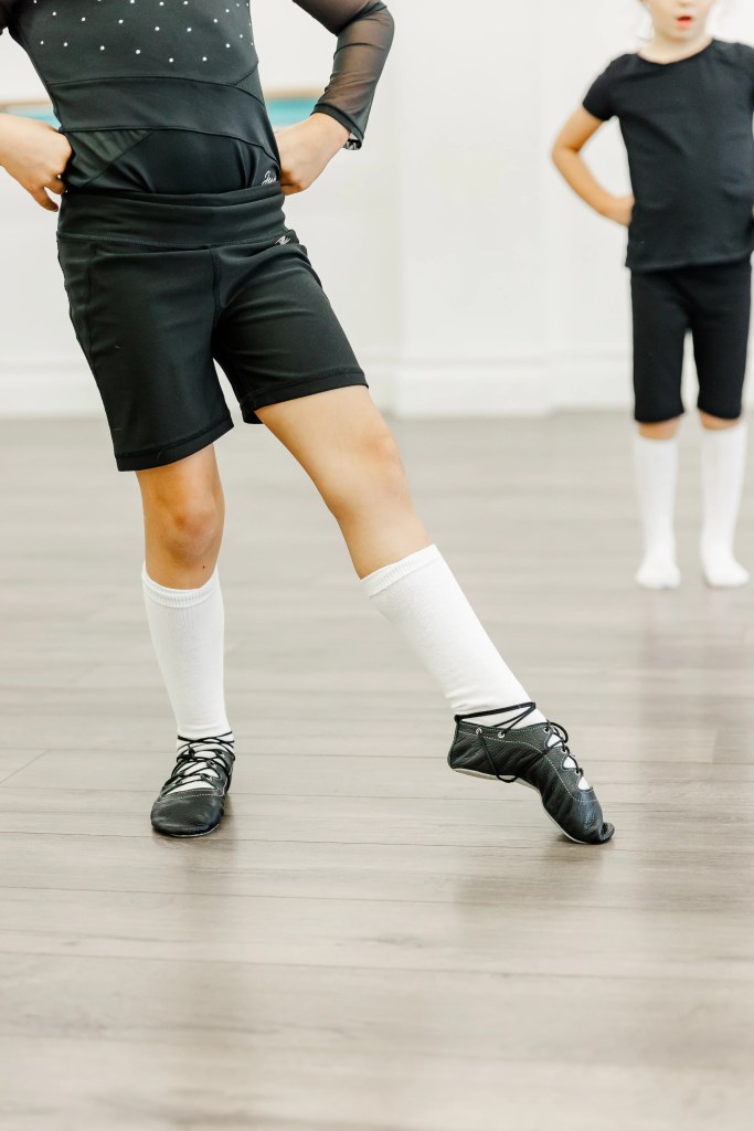 A young dancer demonstrates a pointed foot. She wears all black, white socks, and ghillie dance shoes. Saorsa Studio offers Highland Dance classes for ages 1 to adult in Fredericton, New Brunswick.