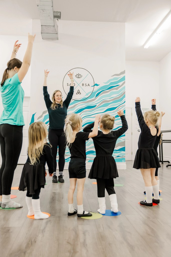 Child highland dancers practice in a studio. Two instructors help the dancers to demonstrate an arm position. Saorsa Studio offers Highland Dance classes for ages 1 to adult in Fredericton, New Brunswick.