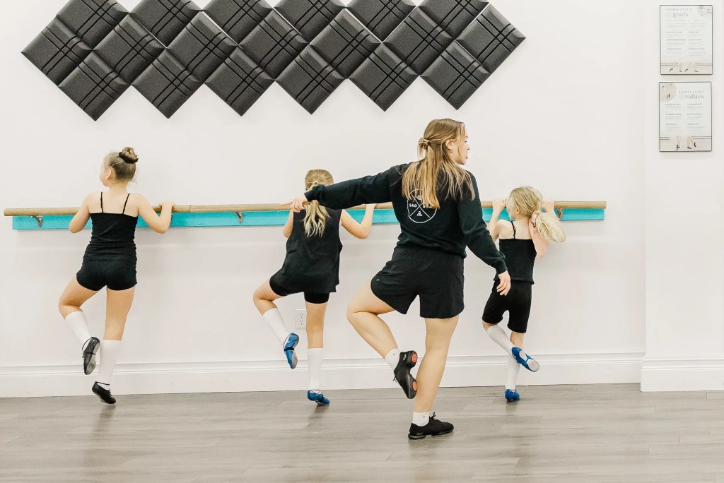 3 dancers hold onto a dance barre. An instructor wearing black coaches them. Saorsa Studio offers Highland Dance classes for ages 1 to adult in Fredericton, New Brunswick.
