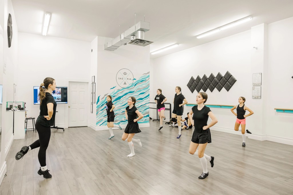 Young highland dancers practice together in a white dance studio. A dance instructor, Nicole Odo, wears black at the front of the class. Saorsa Studio offers Highland Dance classes for ages 1 to adult in Fredericton, New Brunswick.