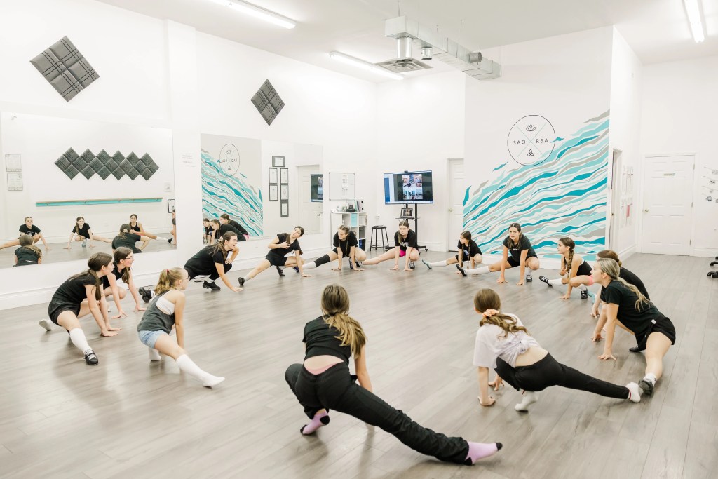 Young highland dancers stretch in a circle in a dance studio. The wall behind them reads "Saorsa". Saorsa Studio offers Highland Dance classes for ages 1 to adult in Fredericton, New Brunswick.