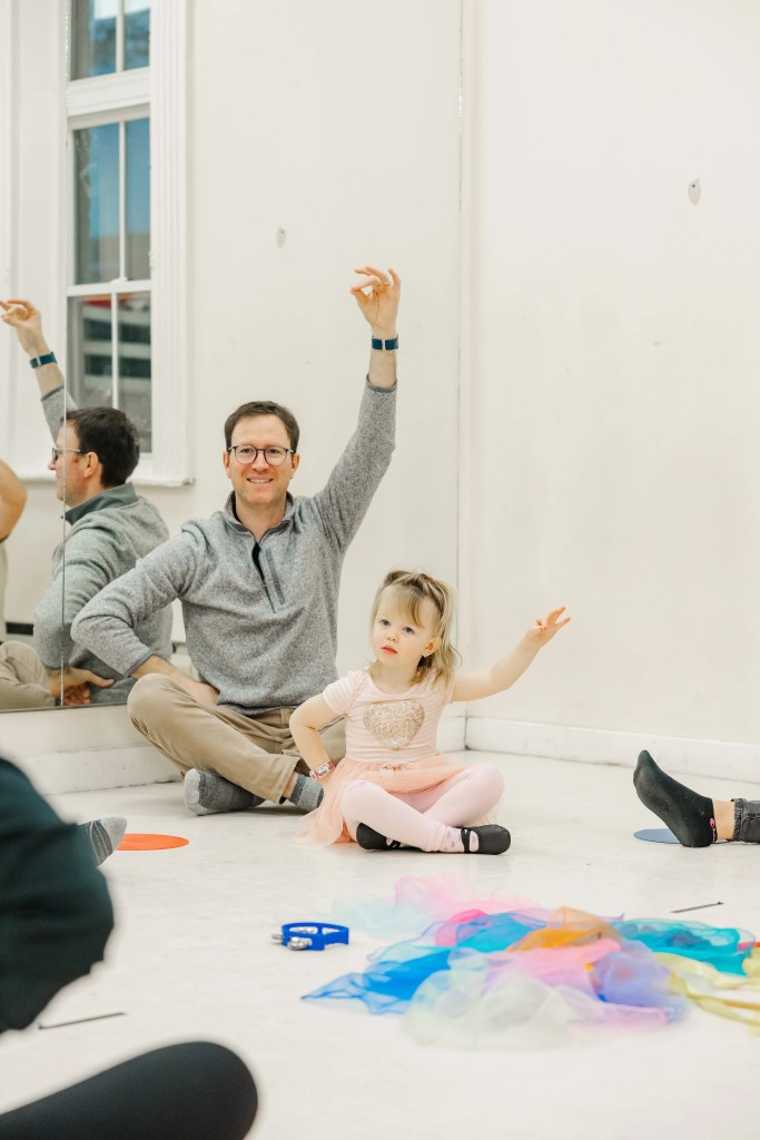 A father and child sit on the ground, demonstrating a highland dance arm pose. Saorsa Studio offers Highland Dance classes for ages 1 to adult in Fredericton, New Brunswick.