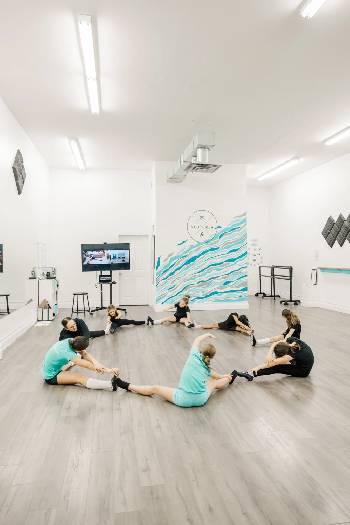 Children wearing black and teal dance clothing highland dance in a white room. They stretch in a circle. Saorsa Studio offers Highland Dance classes for ages 1 to adult in Fredericton, New Brunswick.