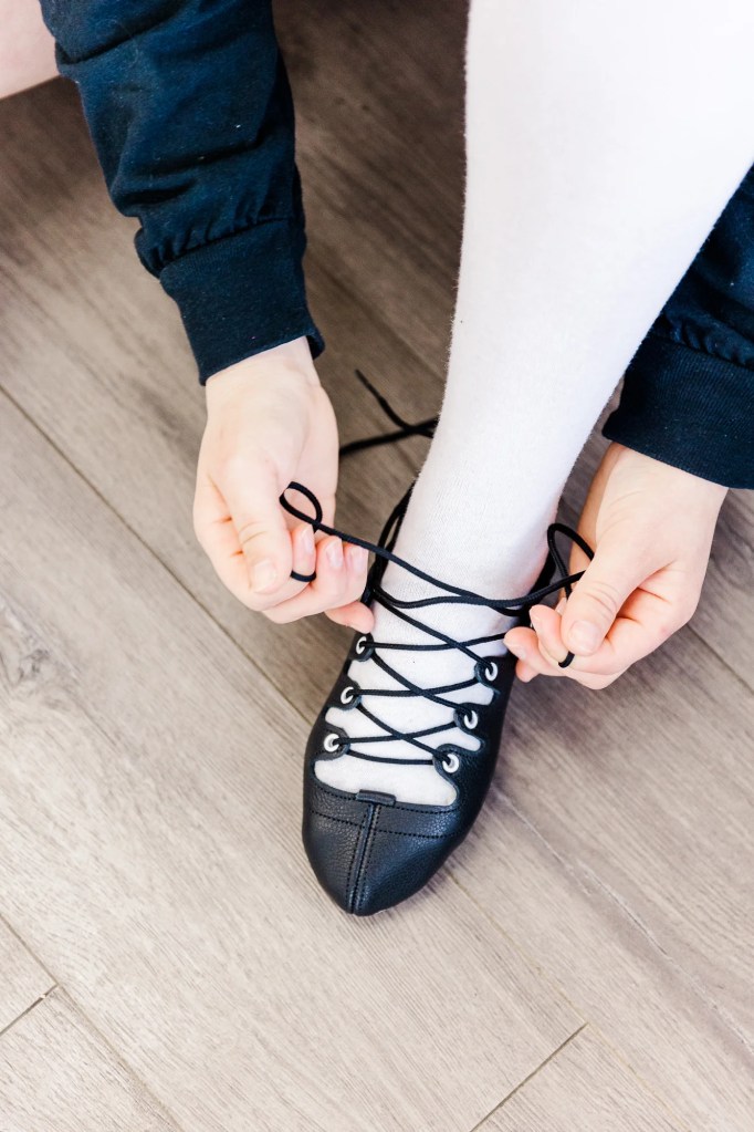 A highland dancer sits on the floor wearing white socks and tying their black leather shoe. Saorsa Studio offers Highland Dance classes for ages 1 to adult in Fredericton, New Brunswick.