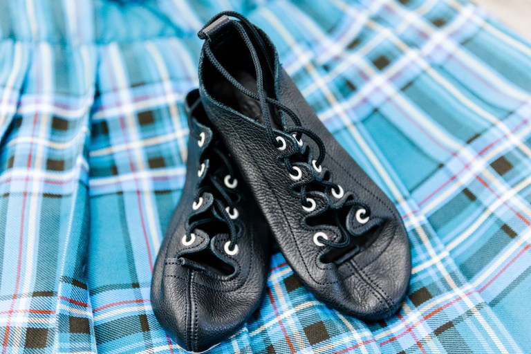 A pair of black leather highland dance ghillies shoes sits on a blue tartan backdrop. Saorsa Studio offers Highland Dance classes for ages 1 to adult in Fredericton, New Brunswick.