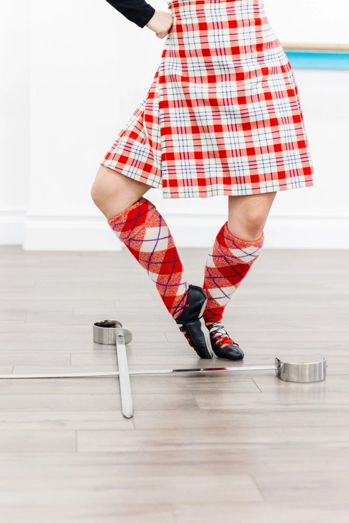 A woman in a red kilt demonstrates a highland dance movement over swords. Saorsa Studio offers Highland Dance classes for ages 1 to adult in Fredericton, New Brunswick.